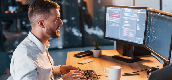 Na imagem um homem está em frente a duas telas de computador digitando códigos de programação, em alusão a um software personalizado e prateleira de prateleira