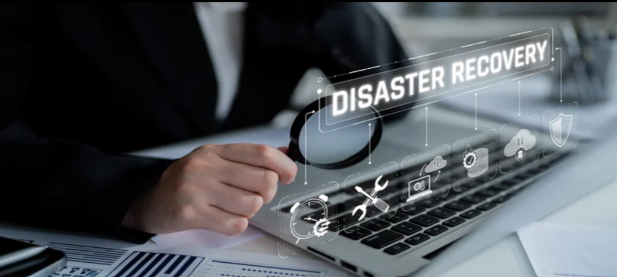 Homem segura uma lupa em frente a notebook com escritos "disaster recovery" e símbolos relacionados desenhados à frente da tela