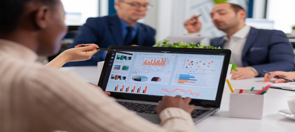 funcionários sentados ao redor de uma mesa de reuniões. Em foco, um notebook aberto com dados expostos em dashboards e um homem de costas, que veste camisa branca de mangas longas, olha direramente para a tela numa análise de consultoria de dados.