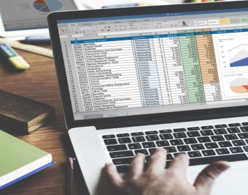 maos humanas digitam em laptop cinza claro com tela focada em planilhas e gráficos de dados, simbolizando uma consultoria de bi