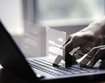 mãos humanas que digitam sobre laptop cinza. Acima da máquina está um elemento gráfico que represanta códigos de IA, representando a atuação de prompt injection