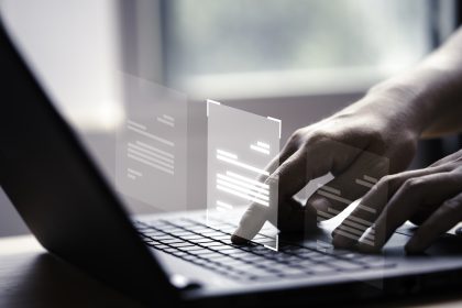 mãos humanas que digitam sobre laptop cinza. Acima da máquina está um elemento gráfico que represanta códigos de IA, representando a atuação de prompt injection