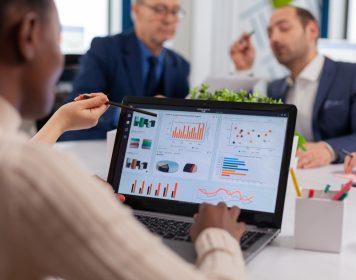 time de master data manegement discutindo ao redor de uma mesa de reunião, com notebook aberto sobre ela demonstrando gráficos de dados a serem gerenciados
