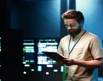 Cientista de dados olha para material em sua mão, enquato ao fundo estão diversos monitores realizando análises de dados