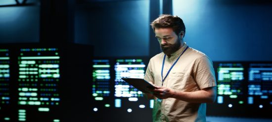 Cientista de dados olha para material em sua mão, enquato ao fundo estão diversos monitores realizando análises de dados