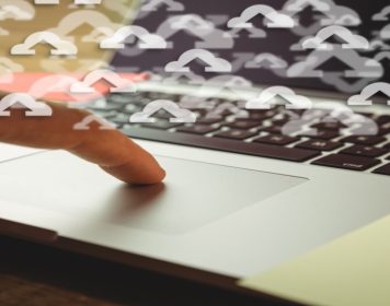 mãos humanas digitando em teclado de noteboo cinza com símbolos de nuvem branco com sinais de seta dentro deles, simbolizando o replatforming