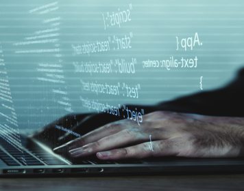 mãos humanas digitando códigos de recodificação em notebook que é cinza e está de costas na imagem. à frente da tela, saem, em branco, códigos de recodificação.