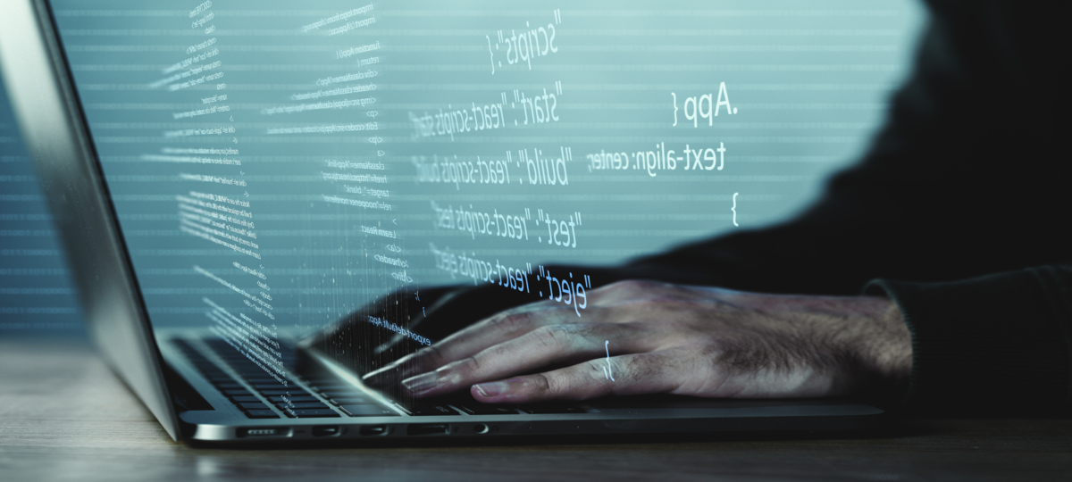mãos humanas digitando códigos de recodificação em notebook que é cinza e está de costas na imagem. à frente da tela, saem, em branco, códigos de recodificação.