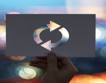 mão humana segurando papel com recorte vazado em formato de "refação", sugerindo o refatoring trabalhando ao longo do conteúdo; Ao fundo, há em desfoque luzes coloridas vindas de máquinas como computadores, o que relaciona a imagem à tecnologia.